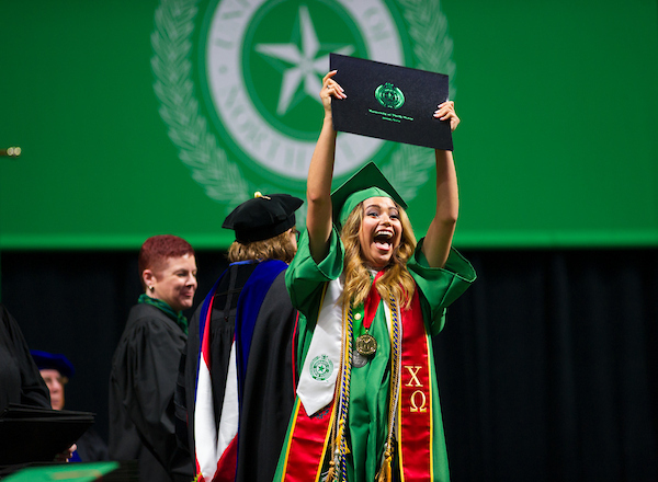 A student in a red stole joyfully holding up her diploma at commencement