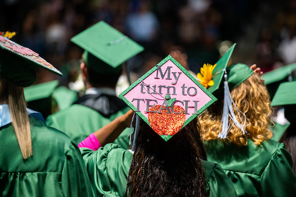 A student at commencement with a cap that says "My turn to teach" over an apple design