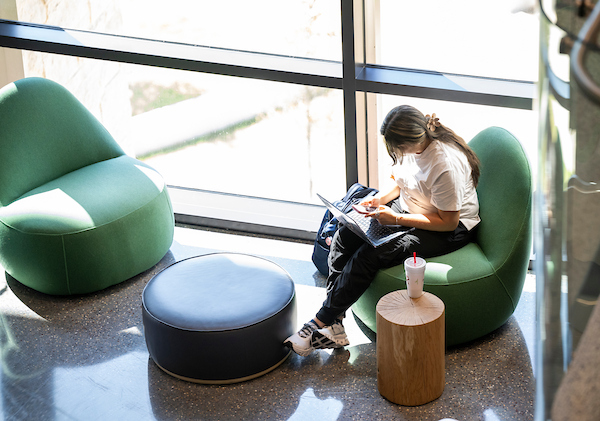 A student sitting in the Frisco atrium works on a phone and laptop