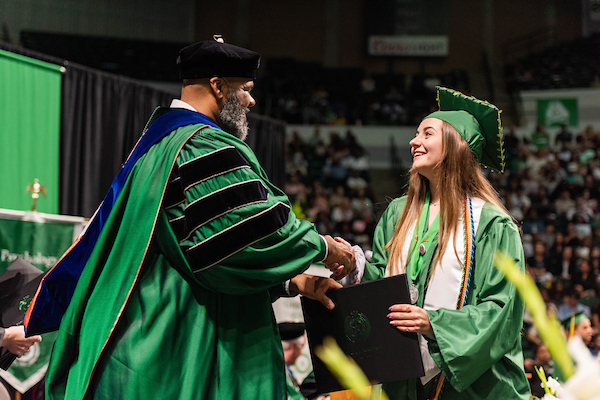 A student in a white honors stole shakes hands with a faculty member and accepts her diploma at commencement