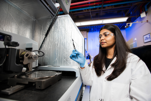 A woman in a labcoat and protective eyewear holds a microchip with tweezers near lab equipment
